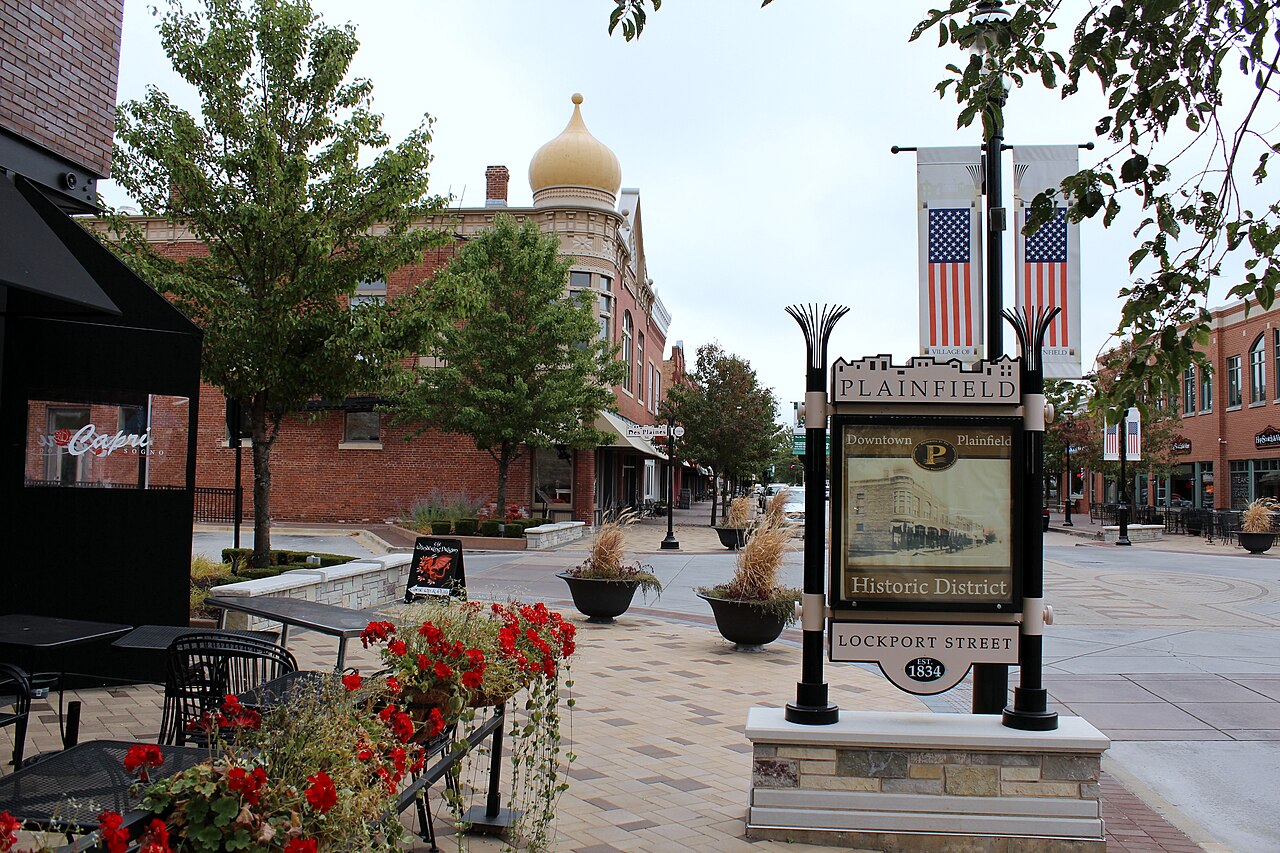 Downtown Plainfield, Illinois, in daylight, with the PLAINFIELD Historic District and Lockport Street sign, brick storefronts, and flower planters.